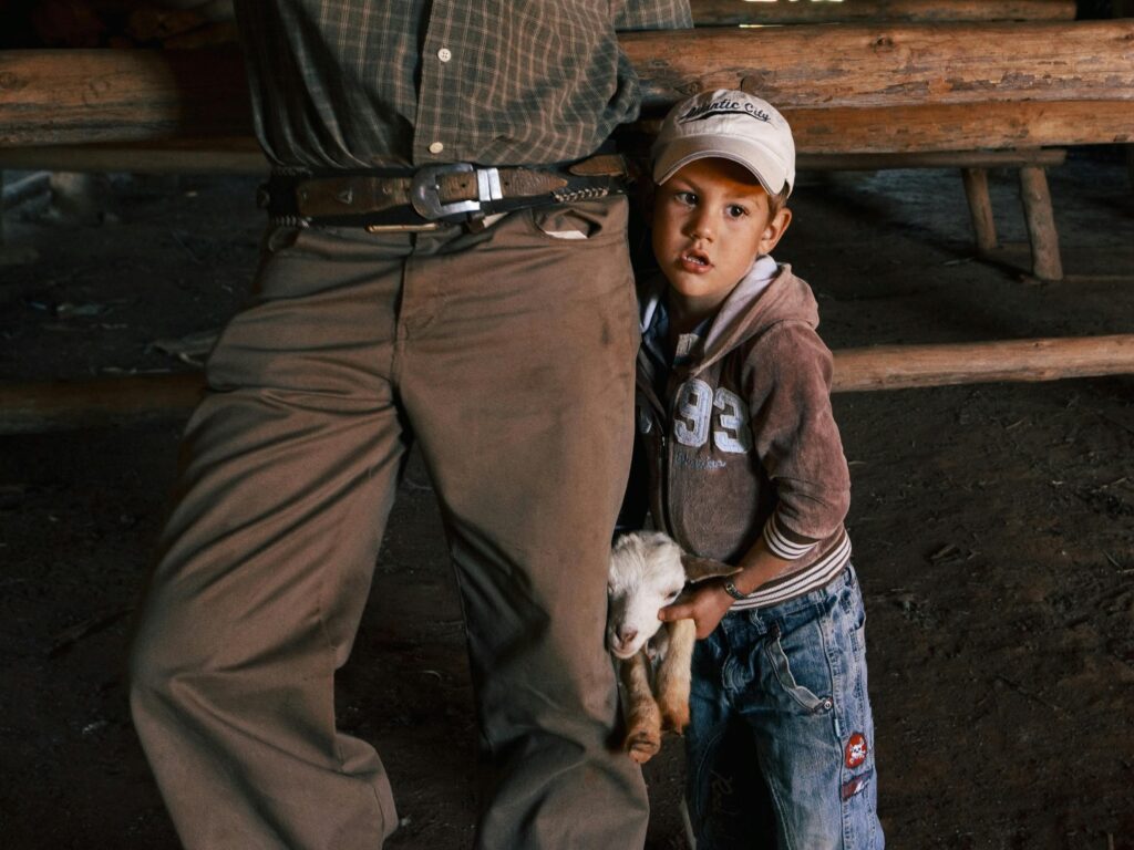 A young boy clings to an adult while holding a baby goat indoors.