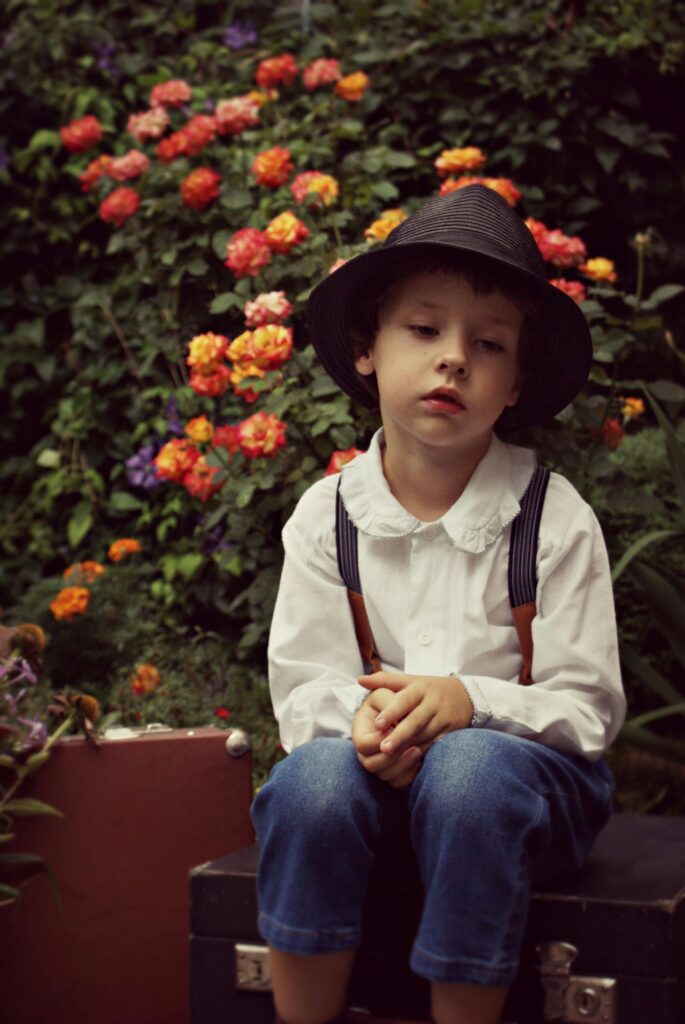 A contemplative boy in vintage attire sits in a garden surrounded by colorful flowers.
