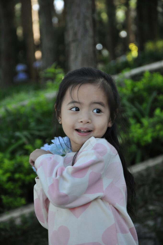 Cute Asian baby girl smiling in an outdoor portrait, surrounded by lush greenery.