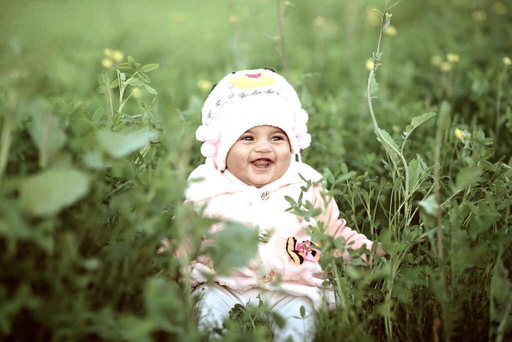 Cute baby in a warm hat laughing amidst green grass in a sunny field.